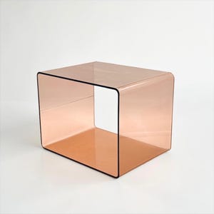 Könnte beinhalten: Ein rechteckiger Beistelltisch aus durchscheinendem, pfirsichfarbenem Acrylmaterial. Das Stück zeichnet sich durch ein durchgehendes, gebogenes Design mit abgerundeten Ecken und einer hohlen Mitte aus, was eine offene, kastenartige Struktur mit glatter Oberfläche ergibt.