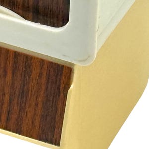 K&ouml;nnte beinhalten: Nahaufnahme einer Ecke einer Holzkiste mit wei&szlig;em Rahmen und goldfarbener Seite. Die Holzmaserung ist im dunkelbraunen Bereich sichtbar. Der wei&szlig;e Rahmen hat eine abgerundete Ecke.