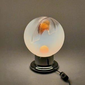 May include: A white frosted glass lamp with a round base and a clear glass globe. The globe has a carved design and a glowing orange light inside. The lamp is on a black base with a cord and switch.