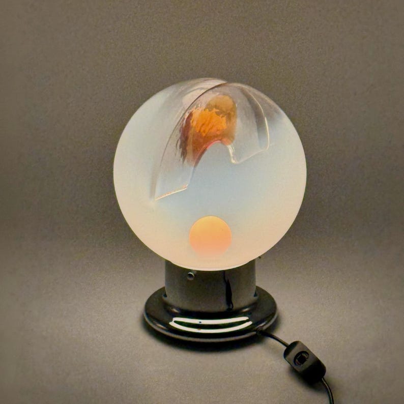 May include: A white frosted glass lamp with a round base and a clear glass globe. The globe has a carved design and a glowing orange light inside. The lamp is on a black base with a cord and switch.