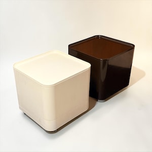 May include: Two square storage containers. One is cream-colored, the other is dark brown. Both have a lid and are on small wheels. The containers are made of a smooth, solid material and are set against a white background.