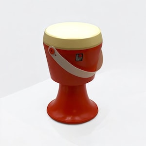 Peut inclure: Un tabouret en plastique rouge et beige en forme de tambour avec une poignée. Le tabouret a une base évasée et un dessus rond. Le dessus est beige et le corps est rouge. La poignée est beige, et la marque "Flair" est imprimée sur le côté.