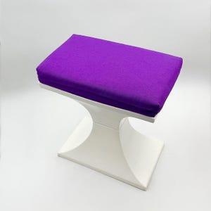May include: A small, white, curved-leg stool with a rectangular, purple cushion. The cushion is a solid, vibrant purple color. The stool's base has a modern, minimalist design.
