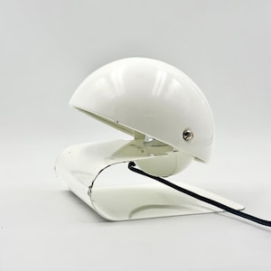 May include: A white, retro-style desk lamp with a curved base and a half-dome shade. The lamp has a glossy finish and a black power cord. The shade is adjustable, and the lamp is designed to sit on a desk or table.
