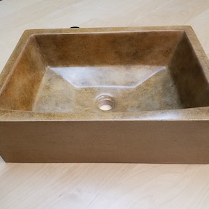 May include: A rectangular concrete vessel sink with a drain hole in the center. The sink is a light brown color with a slightly textured surface.