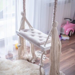 Könnte beinhalten: Eine weiße Holzschaukel mit einem beigefarbenen Kissen und dickem weißem Seil mit Quasten. Die Schaukel hängt von der Decke in einem Raum mit einem weißen Teppich und Holzboden.