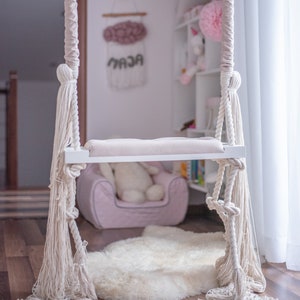 Könnte beinhalten: Eine weiße Schaukel mit einem rosa Kissen hängt an einer weißen Decke in einem Raum mit einem weißen Teppich und einem rosa Sessel. Die Schaukel hat weiße Seilquasten.