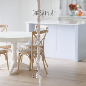 Op de afbeelding: Een witte en beige touw schommel hangend aan het plafond in een kamer met een witte tafel en stoelen. De tekst "OH, SWING!" staat op de muur.