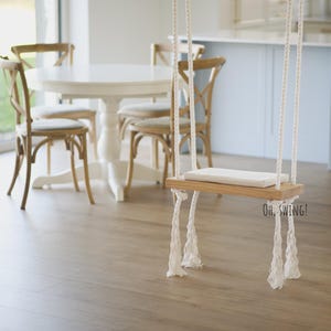 以下が含まれることがあります： 白いテーブルと椅子のある部屋に、白いロープで吊るされた白い木製のブランコがあります。ブランコは木製の座席とロープの端に白いタッセルが付いています。ブランコの真下の床には「OH, SWING!」という文字が印刷されています。