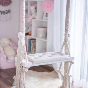 Könnte beinhalten: Eine weiße Holzschaukel mit einem beigefarbenen Kissen und dickem weißem Seil, das von der Decke hängt. Die Schaukel befindet sich in einem Raum mit einem weißen Teppich und einem weißen Bücherregal mit rosa und grauen Dekorationen.