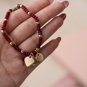 May include: A red beaded bracelet with gold accents and two gold heart charms. The bracelet is on a hand.