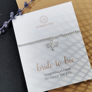 May include: A silver bracelet with a small bee charm. The bracelet is on a white card with the text "bride to bee" and "Congratulations! So happy for you".