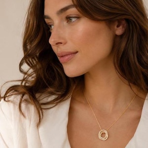 Può includere: Una collana color oro con un ciondolo composto da tre anelli circolari intrecciati. Il ciondolo pende da una sottile catenina color oro. Gli anelli presentano una superficie testurizzata e sono collegati tra loro in un'unica unità circolare.