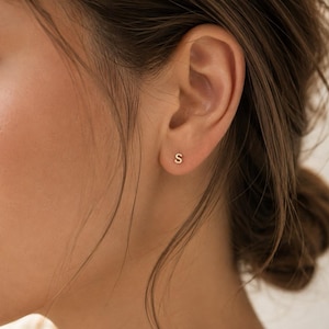 May include: A small, gold-toned stud earring shaped like the capital letter S. The earring is positioned in the centre of the earlobe, featuring a polished metallic finish that reflects light, creating a simple and clean appearance on the skin.