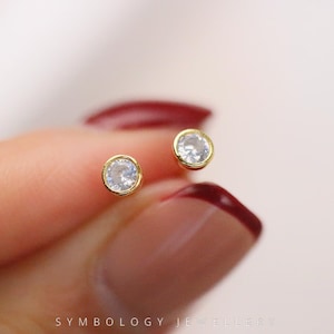 May include: A pair of small, round stud earrings. Each earring features a clear, sparkling gemstone set in a gold-colored bezel. The earrings are held against a blurred background, with the text "SYMBOLOGY JEWELLERY" at the bottom.