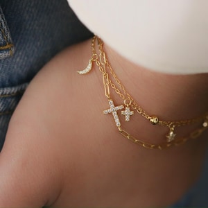 May include: A gold-colored bracelet with multiple strands. The bracelet features charms including a crescent moon, crosses, and stars, all adorned with small, sparkling stones. The jewelry is worn on a wrist, with denim and a white sleeve in the background.