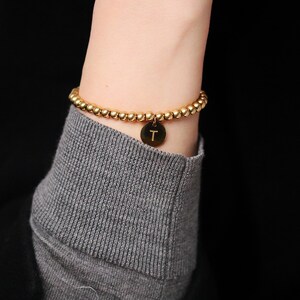 Könnte beinhalten: Ein goldfarbenes Armband mit Perlen und einem goldenen Anhänger mit dem Buchstaben "T".