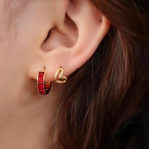 Puede incluir: Primer plano de una oreja adornada con dos pendientes. Uno es un aro dorado con piedras rectangulares rojas, y el otro es un pequeño pendiente dorado en forma de corazón. Los pendientes se destacan sobre la piel y el cabello de la persona.