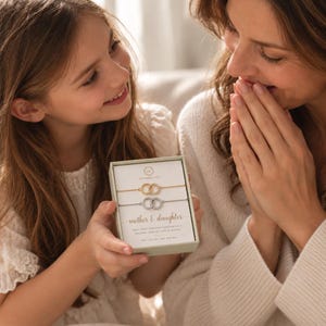 Puede incluir: Una caja de regalo rectangular que contiene dos pulseras delicadas, una dorada y otra plateada, cada una con dos dijes circulares entrelazados. El inserto de la caja muestra el texto "Symbology", "mother & daughter" y texto adicional impreso.