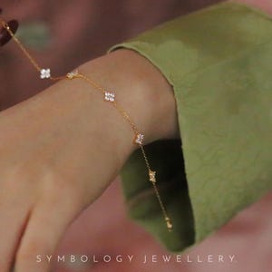 Puede incluir: Una delicada pulsera dorada con abalorios de tréboles de cuatro hojas y pequeños detalles brillantes. La pulsera se lleva en una muñeca, con una manga verde de fondo. El texto "SYMBOLOLOGY JEWELLERY" está en la parte inferior.