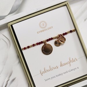 May include: A gold bracelet with red beads and charms in a white box with the text "Symbology" and "fabulous daughter Love you today and always x".