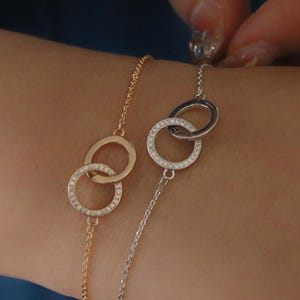 Peut inclure: Deux bracelets délicats, l'un doré et l'autre argenté, chacun avec des cercles entrelacés. Les cercles sont ornés de petites pierres scintillantes. Les bracelets sont présentés sur le poignet d'une personne.