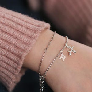 Puede incluir: Una pulsera plateada con dos cadenas y dijes. Uno es la letra "R", y el otro es un perro globo. La pulsera se lleva en la muñeca, con un suéter rosa en el fondo.