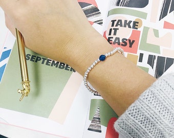 Peut inclure: Un bracelet en argent composé de perles avec une seule perle bleue. Le bracelet est porté au poignet d'une personne et l'arrière-plan est une page colorée d'un agenda avec les mots "Take it easy" et "September".