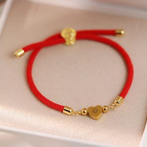 May include: A red cord bracelet with gold-colored accents and a heart-shaped charm. The bracelet is presented in a white box with a soft, neutral-toned interior. The heart charm has a sunburst design.
