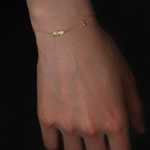 Op de afbeelding: Een delicate gouden kettingarmband met een klein bedeltje met een cluster van kleine, heldere steentjes. Een enkel helder steentje hangt aan de ketting.