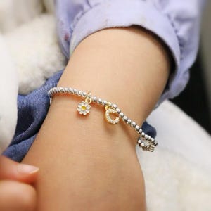 Op de afbeelding: Een zilveren kralenarmband met twee gouden bedels, een madeliefje en een letter "C" bezet met kleine heldere steentjes. De armband wordt om een pols gedragen. Op de achtergrond een wit knuffeldier en een blauw overhemd.