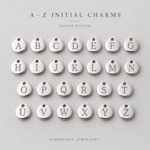 Könnte beinhalten: Eine Sammlung von versilberten runden Anhängern, die jeweils mit einem einzelnen Großbuchstaben von A bis Z graviert sind. Der Text A-Z INITIAL CHARMS und SYMBOLOGY JEWELLERY steht über und unter dem Raster aus sechsundzwanzig Metallanhängern.