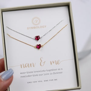 Może przedstawiać: Dwa delikatne bransoletki z czerwonymi zawieszkami w kształcie serca, prezentowane w białym pudełku z tekstem "nan & me wear these bracelets together as a reminder that our love is forever ONE FOR YOU, ONE FOR ME".