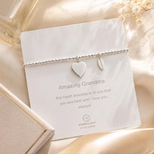 Pode incluir: Uma pulseira de contas prateadas com um pingente em forma de coração e uma pequena etiqueta retangular. A pulseira é apresentada num cartão branco com o texto Amazing Grandma, this heart bracelet is to say that you are best and I love you, always!
