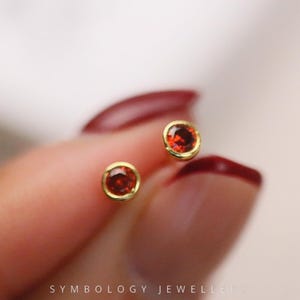 May include: A pair of small, round stud earrings. Each earring has a vibrant, round, red gemstone set in a gold-coloured bezel. The earrings are displayed against a blurred background, with the text "SYMBOLGY JEWELLER" at the bottom.