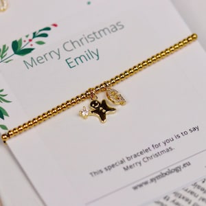 Könnte beinhalten: Goldenes Perlenarmband mit drei Anhängern: einem Lebkuchenmann, einem Stern und einem Blatt. Das Armband liegt auf einer Karte mit der Aufschrift "Merry Christmas Emily". Die Karte hat ein festliches grün-rotes Blumenmuster.