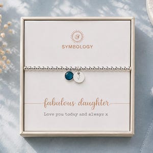 Puede incluir: Una pulsera elástica de cuentas plateadas con un pequeño dije de piedra azul oscuro y un disco plateado grabado con la letra J. La pulsera se presenta en una caja cuadrada con el texto Symbology, fabulous daughter, y Love you today and always x.