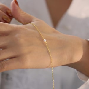 May include: A delicate gold bracelet with a small, round, white pearl accent. The bracelet is on a slender chain and is being worn on a wrist. The background is a soft, neutral colour.