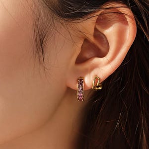 May include: A gold hoop earring featuring a row of small, square-cut purple gemstones and a second smaller gold hoop earring shaped like a heart, both worn in the earlobe. The jewellery is crafted from a polished gold-tone metal material.