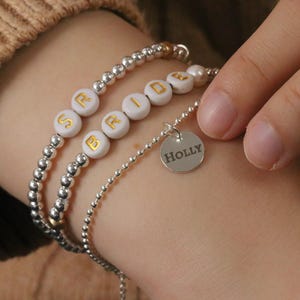 May include: A silver beaded bracelet with the word "BRIDE" spelled out in white beads with gold lettering. A smaller silver chain bracelet with a silver charm that says "HOLLY" is also on the wrist.