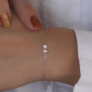 Puede incluir: Una delicada pulsera de plata con un diseño en forma de lazo con dos gemas claras y brillantes. La pulsera se muestra en una muñeca, con una camisa blanca en el fondo. Las gemas miden aproximadamente 0,6 cm.
