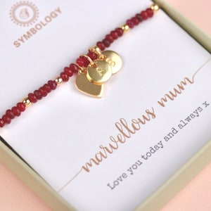 Puede incluir: Un brazalete de cuentas rojas con dijes de corazón y letras doradas. El brazalete está en una caja con una tarjeta que dice "marvelous mum" y "Love you today and always x".