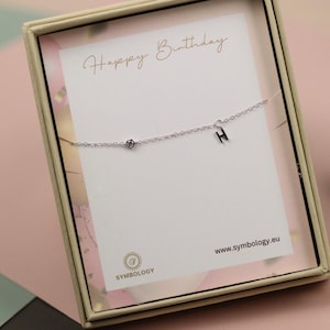 Könnte beinhalten: Eine silberne Halskette mit einem kleinen Diamanten und dem Buchstaben "H" wird in einer Geschenkbox präsentiert. Die Box hat einen goldenen Rahmen und eine weiße Karte mit den Worten "Happy Birthday" in Schreibschrift. Die Website www.symbology.eu ist ebenfalls sichtbar.