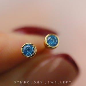 May include: Close-up of two gold stud earrings, each featuring a round, light blue gemstone. The earrings are set in a gold bezel setting. The background is a soft, blurred pink.