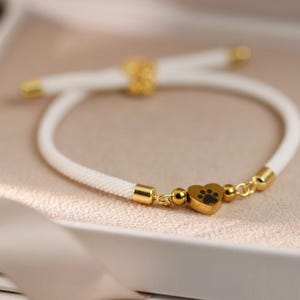 Könnte beinhalten: Weißes Seilarmband mit goldfarbenen Akzenten und einem herzförmigen Anhänger mit schwarzem Pfotenabdruck. Das Armband wird in einer weißen Box mit Satinband präsentiert.