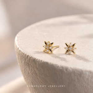 May include: A pair of gold stud earrings featuring a star-shaped design with four pointed petals and a central cluster of small clear stones. The earrings have a polished finish and are marked with the text SYMBOLOGY JEWELLERY at the bottom.