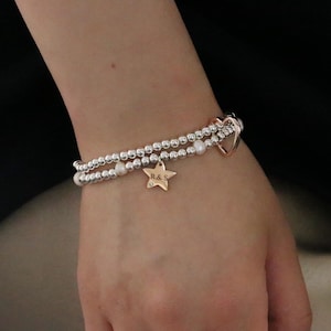 May include: A silver beaded bracelet set with a gold star charm and a gold heart-shaped charm. The star charm has the initials "R & S" engraved on it. The bracelet also features small white pearls.