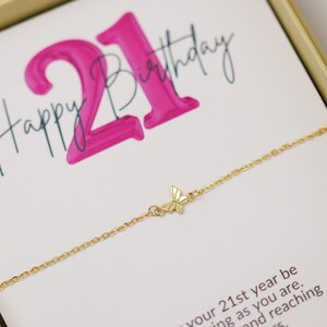 May include: A gold butterfly charm bracelet on a white card with the words "Happy 21st Birthday" in black script and pink block letters. The bracelet is inside a gold box.