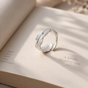 May include: A silver-toned ring shaped like a curved feather with detailed etched lines along the surface. The ring is open-ended and rests on an open book page featuring the printed text Small steps make a big difference and A kinder brighter you.
