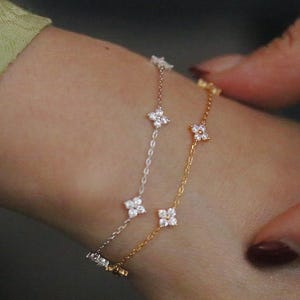 May include: Three delicate bracelets with floral designs. One is gold, one is silver, and the third is rose gold. Each bracelet features small, sparkling flower-shaped accents. The bracelets are worn on a wrist.